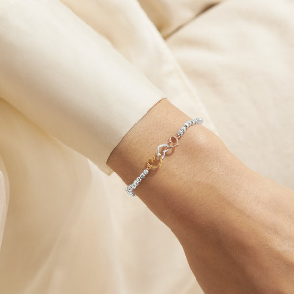 Joma Jewellery beaded bracelet with three interlinked hearts in silver, gold and rose gold presented on a blush pink sentiment card.